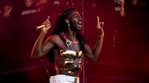 Lil Nas X sostiene un micrófono y no usa camisa, excepto platos de oro y pantalones blancos