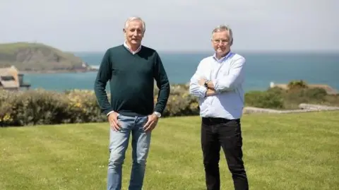 Two men wearing jeans and shirts standing on a lawn in the sunshine against a backdrop of blue sea and picturesque cliffs.