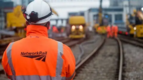A man wearing an orange high visibility jacket with Network Rail written on it. He is wearing a helmet looking at a railway line with various vehicles in the background and workers.