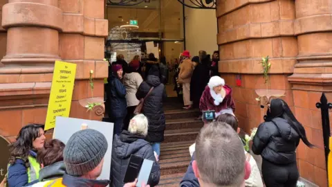 Para pengunjuk rasa Johnlawrence berkumpul di tangga di pintu masuk ke kantor Greater Manchester Gabungan Oxford Street. Beberapa dapat dilihat dekat dengan pintu kaca yang mengarah ke dalam markas. 