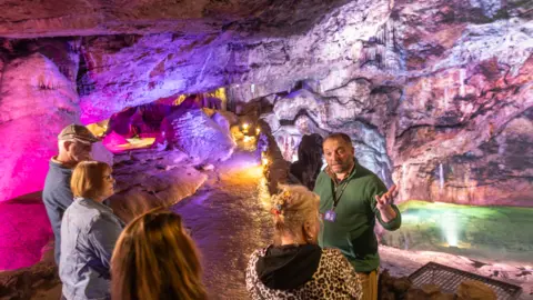 Wookey Hole Caves Four visitors and a male guide stand in a cave which is brightly lit with a purple hue. The man is gesticulating with his left hand and is wearing a green jumper and a lanyard. The path weaves away into the back of the cave with yellow lowlights lighting the way