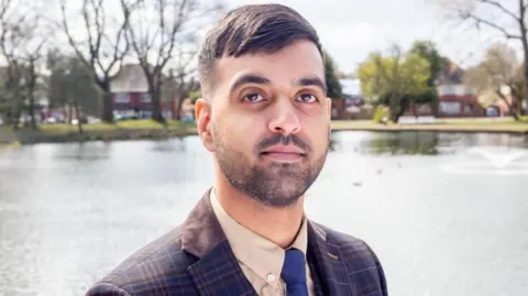 A man with short black hair and a short beard, wearing a suit jacket, shirt and blue tie, stands looking at the camera. A lake is behind him with trees and houses visible on the other side.