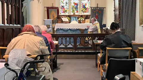 Qays Najm/BBC The Reverend Jeremy Lind during a church service that he is leading. Churchgoers sit in pews praying as the priest speaks at the front. 