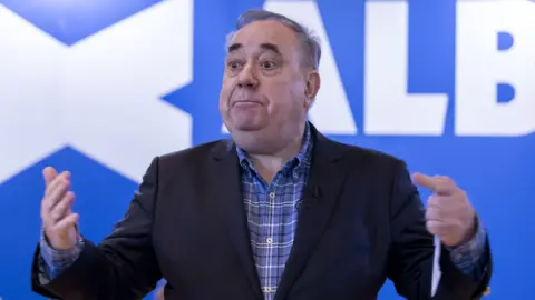 PA Media Alex Salmond standing in front of a blue background with the Alba Party logo on it.