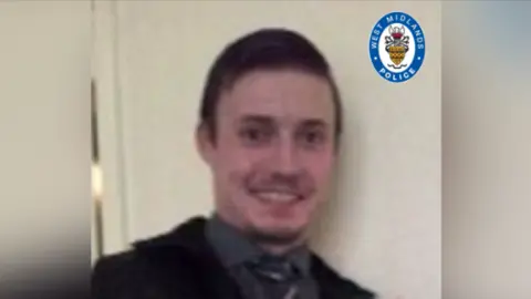 Daniel Lewis is dressed in a black suit and dark grey shirt with a dark striped tie. He is smiling at the camera and has a small goatee and short hair