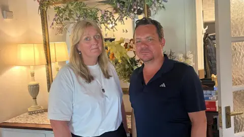 A women wearing a white t-shirt stood with a man wearing a blue polo shirt. They are both looking at the camera. 