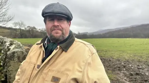 Chris Armstrong, a bearded man wearing a grey wool cap, a beige jacket and a green scarf. He is standing next to a dry stone wall and behind him is a field backing onto woodland, with hills in the distance under a grey sky.