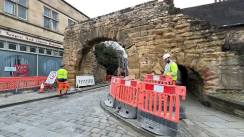 A roman bricked archway. The sandy coloured bricks are chipped in places. Green moss covers patches of the monument. Two people in high vis are standing near the archway. Red plastic fences have been put in place in front of it.