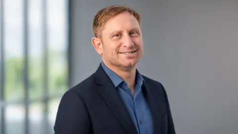 Systal Technology Solutions Man with ginger hair and a beard smiling at the camera. He is wearing a blue shirt and a dark blue jacket. Behind him is the interior of an office building.
