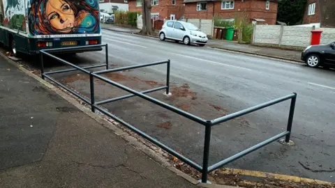 BBC Grey tubular metal fencing, divided into two bays, sited next to a pavement and opening onto the roadway