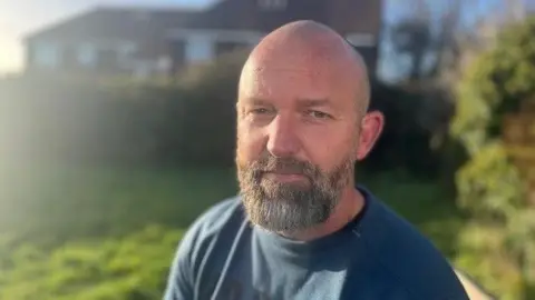A bald man with a beard is looking directly at the camera. He is wearing a grey sweatshirt and there is greenery, slightly out of focus, behind him