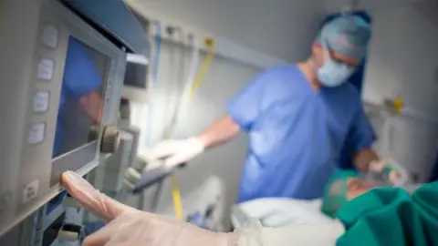 Getty Images A gloved hand presses buttons on a medical monitoring machine in an operating theatre. In the background, a patient lies on a hospital bed covered with green surgical drapes while a healthcare worker in blue scrubs, a surgical cap, mask and gloves stands beside the bed.