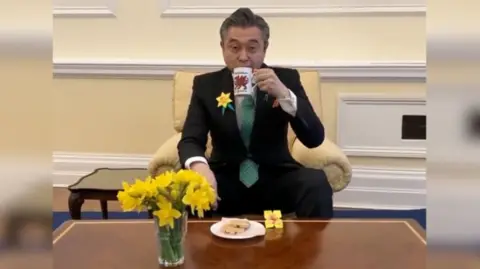 The Japanese ambassador is sat in a cream-coloured armchair. He is wearing a blue suit with a green tie, and is sipping tea from a white mug with a red dragon on it. In front of him is a brown table, which has a vase of daffodils, a paper origami daffodil and a plate of Welsh cakes