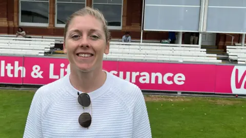 Heather Knight looking towards the camera in a white jumper with sunglasses hooked on the neck.