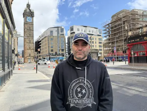 A person wearing a dark hoodie and baseball cap stands on a pavement in central Glasgow near a junction with traffic cones and roadworks, with a clock tower and surrounding buildings visible in the background, including shop windows displaying signage.