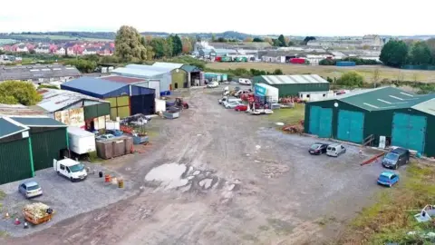An aerial photo of a collection of green industrial buildings around a hardstanding car park. Some of the buildings are patched up or rusty.