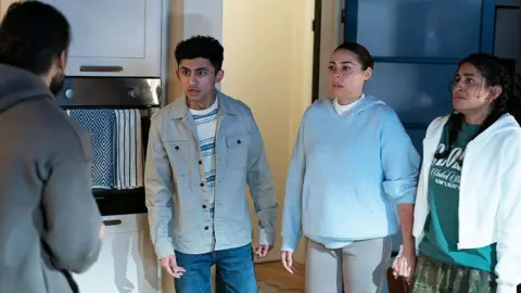 BBC/Jack Barnes/Kieron McCarron A still showing Ravi on the edge of the scene, with family members Nugget, Priya and Avani. They are standing in the kitchen speaking emotionally to Ravi.