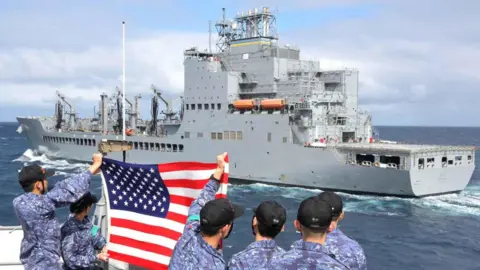 Reuters Crewmembers wave an American flag from another boat on the ocean as the US Navy oiler USNS Harvey Milk