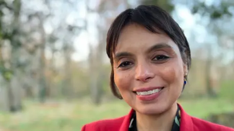 Dr Emma Egging, who is smiling and has short black hair, stands in front of a blurred background of trees and greenery. 