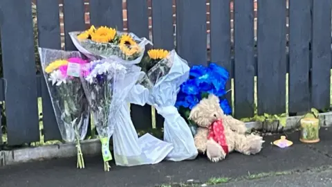 BBC A teddy bear and bouquets of flowers are left by a fence in front of the house in Salford where two bodies were found on Monday