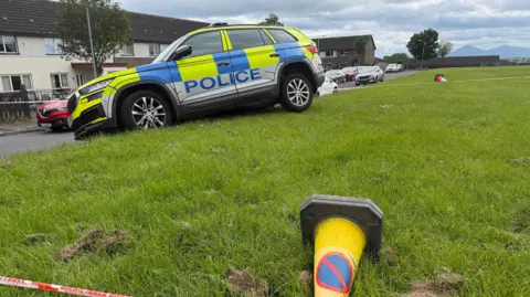 Pacemaker A stolen police car lies abandoned on a grassy bank in the middle of a housing estate. A police traffic cone and a piece of plastic police cordon tape are partially visible in the foreground.