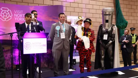 Getty Images Rishi Sunak is standing on stage in front of a bright purple backdrop with the slogan visit North Yorkshire. He is on the left of the photograph, giving a speech at a podium, with other candidates lined up to his right. Count Binface is standing on the right of the stage in his costume.