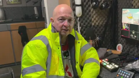 The Phobic Flyer A man sat in front of a desk, leaning on a table with headphones and equipment on it. He is wearing a high-vis jacket.
