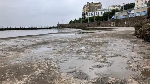 BBC Weston Marine Lake's structure without any water in. There is a rough concrete surface with potholes full of water on a walking around a lake.
