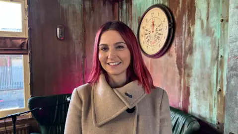 Dale Baxter / BBC Melanie Onn. She is smiling at the camera. She has dark pink hair and is wearing a beige coat. 