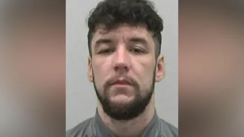 Northumbria Police Mugshot of Hulse. He has short thick black curly hair and a black beard with spots on his neck