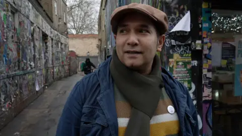 A man wearing a flat cap, striped wool jumper, scarf and jacket looks away from camera with an urban background of graffiti and peeling posters  