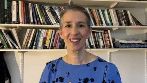 A woman smiling into the camera with a blue top on that has black flowers on it. 