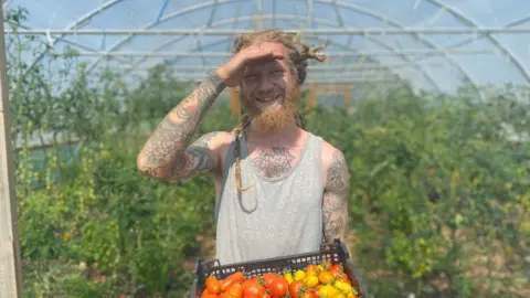 Shem/Amary Farm Will Davis has his hand to his face to shield his eyes from the sun. He is wearing a singlet vest and holding a tray of red, yellow and orange tomatoes. He is standing in a polytunnel with tomato plants growing in it.