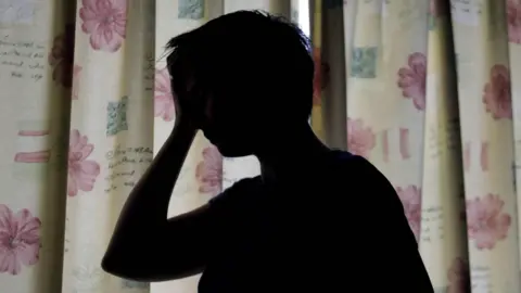 The silhouette of a woman in front of some curtains. She is holding her hand against her face. 