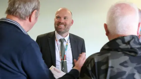 Tom Hardwick is standing in between two people and the backs of their heads are visible. He is is wearing a suit jacket and lanyard and smiling broadly. 