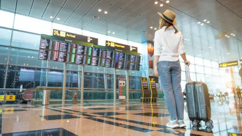 A woman looks at a departures board in an airport. She is dressed in summer clothes