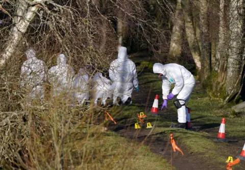 PA Scenes of crime officers in white outfits examine an area of dirt track. Small flags and markers are dotted around the scene