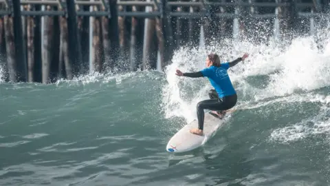 Sean Evans/ISA Surfing Melissa Reid is surfing a wave with her arms stretched out and her knees bent as the surfboard moves down the face of a wave. She is wearing a Team England T-shirt over her wetsuit. Behind her is the pier.