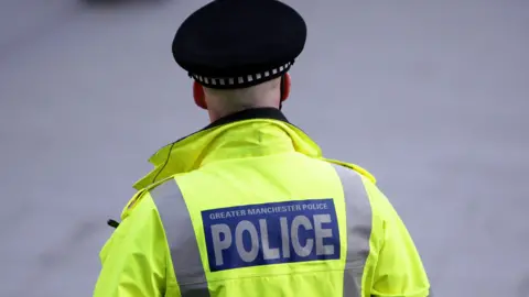 PA Media Generic image of the back of an unidentifiable policeman, who wears a black cap and yellow hi-vis jacket with blue logo saying Greater Manchester Police