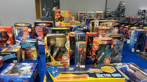 Molly Smith/BBC A collection of Doctor Who toys laid out on a table with a royal blue surface. Everything is in its own box: the toys include a number of different sonic screwdrivers, a mini talking dalek and a mini tardis.