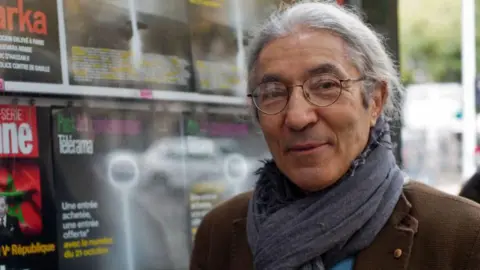 Franco-Algerian writer Boualem Sansal poses in a brown jacket and grey scarf beside a newspaper kiosk in Paris in 2015