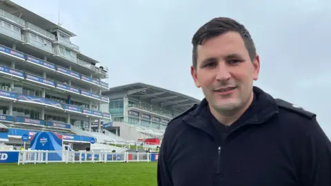 Julia Gregory / BBC A man wearing a black zip up top stood in front of a racecourse grandstand.