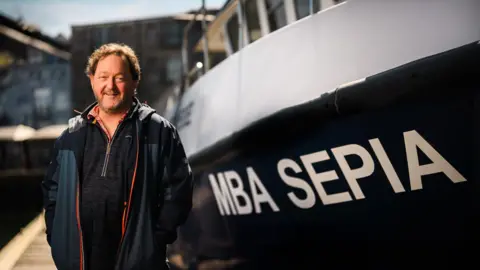 Dr Bryce Stewart wearing a rain coat and fleece is stood next to a yacht that has the lettering MBA Sepia on the side. 