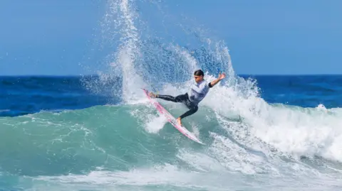 Boardmasters Festival / Jason Feast A surfer in a black wetsuit and white rash vest rides the crest of a wave on a pink surfboard on a sunny day.