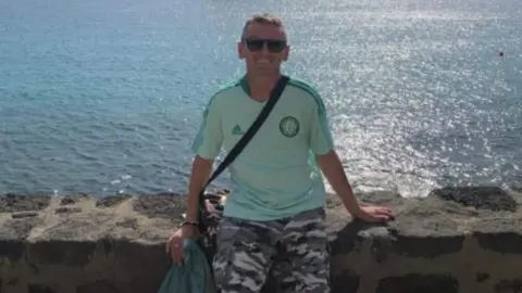 Family handout A man sits on a wall with the sea behind him. He is wearing a pale green Celtic away top, shorts and holding a jacket in one hand, while smiling directly into camera. 