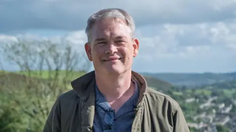 Welsh Labour Richard Tunnicliffe standing on a hillside above Caerphilly on a sunny day. The blurred background includes a tree with no leaves on it to the left of frame. He is clean shaven and wearing a khaki coloured jacket. 