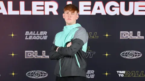 PA Media Angry Ginge stands, arms folded, in front of a Baller League hoarding with his arms folded. He wears a tracksuit top with black, grey and aqua-coloured panels and a determined look on his face.
