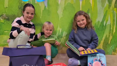 Darlington Council Three children of different ages are sat on the floor, some holding books. They are in a colourful room with a wall that has some artwork on it behind them. They are all smiling at the camera.