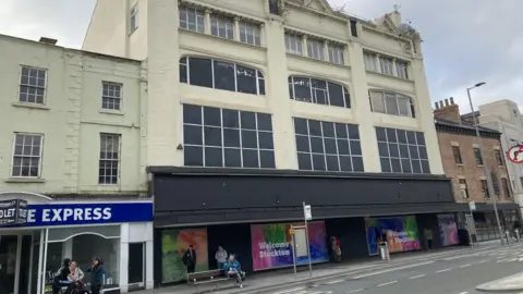 The former Debenhams department store in Stockton. It is a multi-storey building, painted cream with the majority of its windows blacked out. The lower area is painted black and is adorned with signs reading "Welcome to Stockton". Someone is sitting on a bench on the pavement, while three people stand chatting on the street nearby.