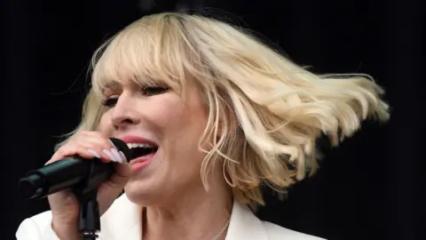 PA Media Natasha Bedingfield sings into a microphone, she has a blonde bob and is wearing a white suit jacket